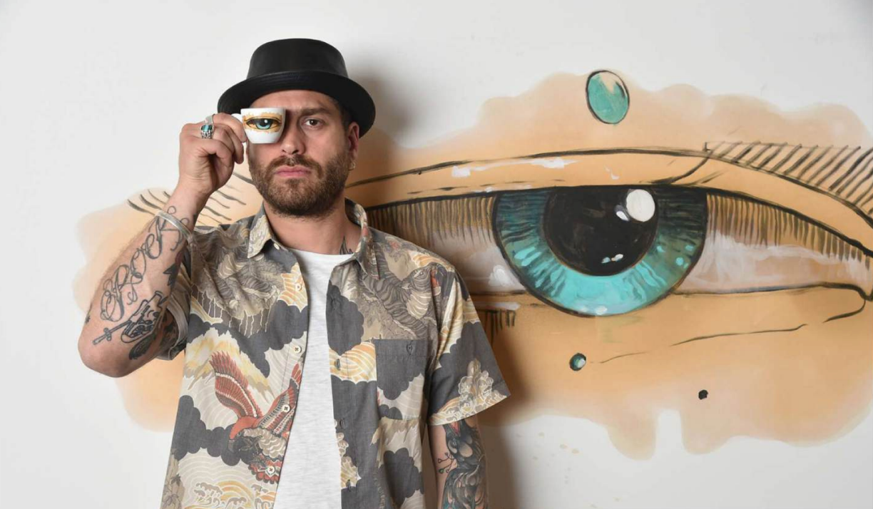 Max Petrone con una tazzina Coffee Drawings davanti al suo murale con l'occhio