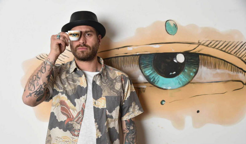 Max Petrone con una tazzina Coffee Drawings davanti al suo murale con l'occhio